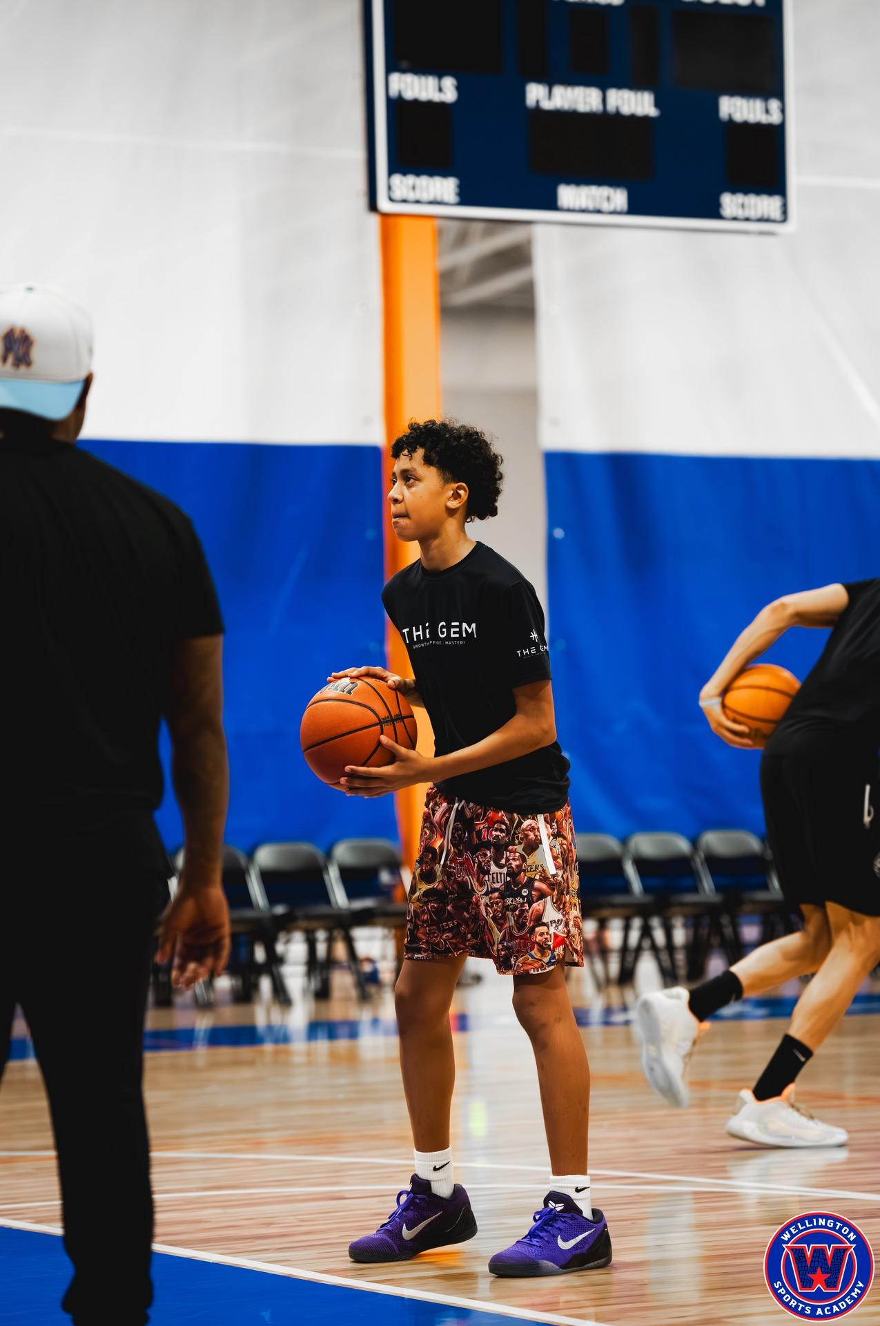 Young player in black "The Gem" shirt holding a basketball mid-drill, coach partially visible