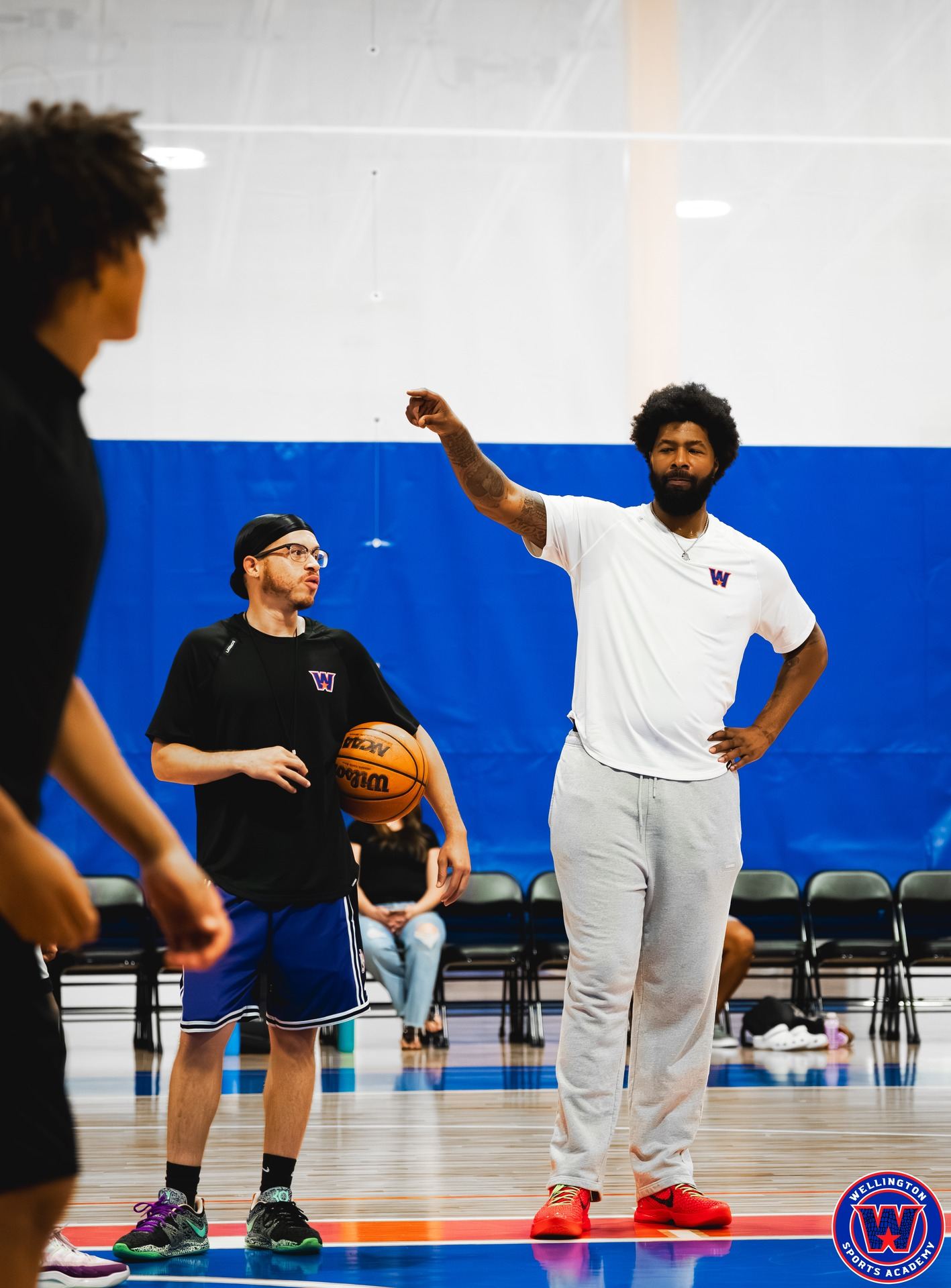 Two coaches — one pointing up, one holding a ball — directing players during a drill