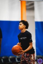 Close-up portrait of young player in black "The Gem" shirt holding a basketball, looking up